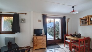 A room with a wooden dresser, a television, a dining table with chairs, a window with curtains, and a door leading outside.