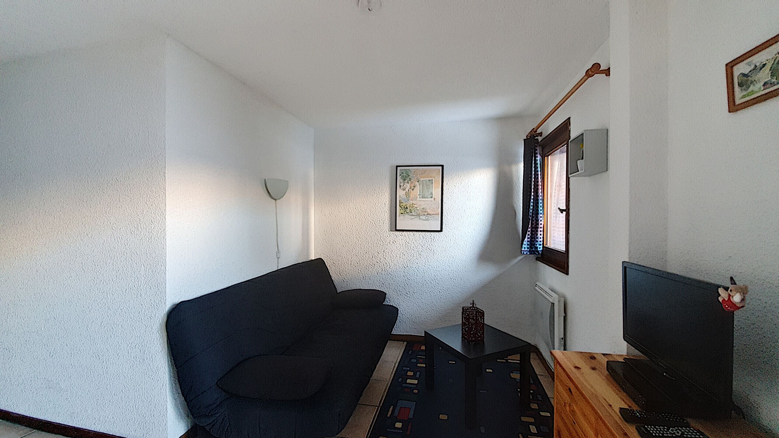 Une petite pièce, peu meublée, avec un canapé sombre, une petite table sur laquelle se trouve une télécommande, un téléviseur et un tableau encadré accroché au mur.
