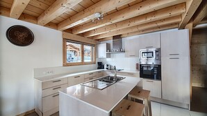 Une cuisine moderne avec des poutres en bois, un grand îlot central, des appareils électroménagers en acier inoxydable et une vue sur l’extérieur par une fenêtre.