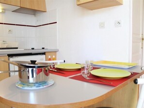 Una cocina con baldosas blancas, armarios de madera, una estufa y un pequeño comedor con un tapete rojo y platos amarillos.