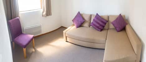 A room with a beige sofa, a purple chair, and a window with curtains.