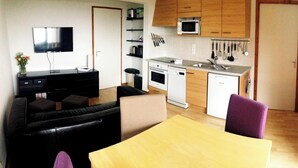 Un espace de vie compact comprenant un canapé, une table à manger, un coin cuisine avec un four à micro-ondes et un évier, ainsi qu’un téléviseur fixé au mur.
