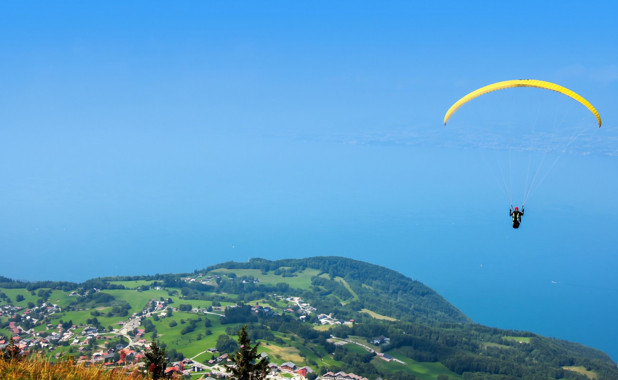 Uma pessoa praticando parapente sobre uma paisagem rural com casas e vegetação abaixo.