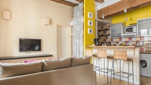 A living room with a sofa, a television, and a kitchen area with a microwave, a coffee maker, and a washing machine.