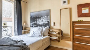A hotel room with a bed, a large mirror, a wooden dresser, and a view of a cityscape through the window.