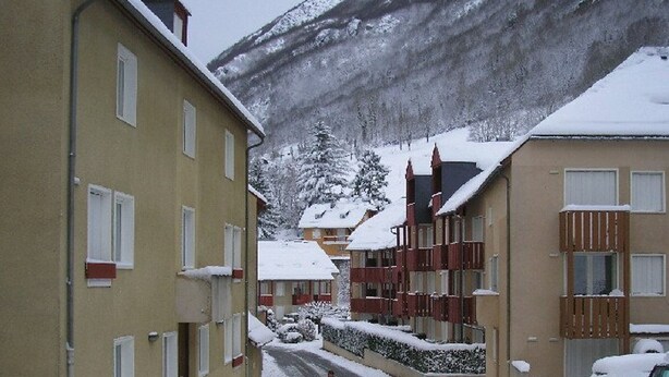 A snow-covered street with buildings on either side, including a multi-story building with balconies and a single-story building with a sloped roof. - Studio cabin in Esquièze-Sère for 6 with balcony, mountain view and parking (Esquièze-Sère)
