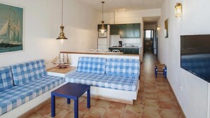 Una sala de estar con un juego de sofá, una mesa de centro y un televisor. Se observa una cocina al fondo.