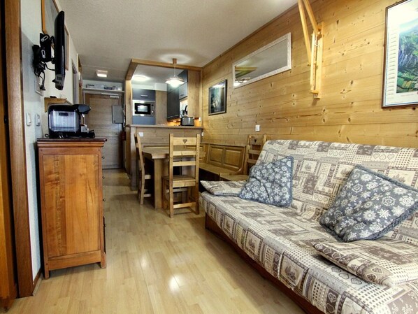 Interior acolhedor de uma cabana, com piso de madeira, um sofá com almofadas estampadas, um armário de madeira e uma kitchenette ao fundo.