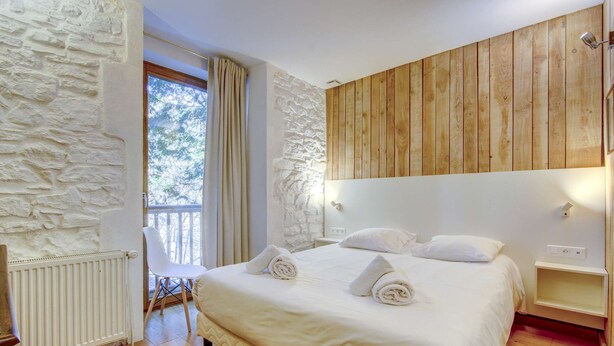 Camera da letto con parete in pietra, testiera in legno intarsiata, un letto con lenzuola bianche e vista su alberi attraverso una finestra.