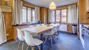 Una stanza con rivestimenti in legno, un grande tavolo da pranzo, sedie bianche, un lampadario e un angolo cottura con forno e frigorifero.