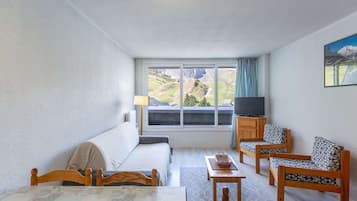 A room with a bed, a sofa, a table with a tray of fruit, a television, and a view of mountains through a window.
