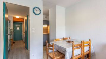 A dining area with a table and chairs, a clock on the wall, and a kitchen in the background.