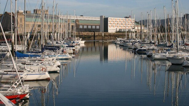 A marina with numerous boats docked, a large building in the background, and calm water reflecting the boats.