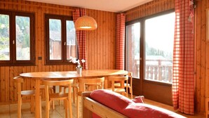 Un interior de madera con una mesa redonda y sillas, un banco con cojines rojos y ventanas amplias con cortinas.
