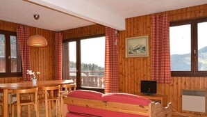 Una habitación de madera con una cama, una mesa de comedor con sillas, un televisor y una ventana que ofrece vistas a las montañas.