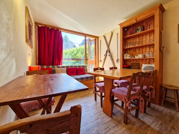 A cozy dining area with a wooden table and chairs, a sofa with red cushions, and a wooden shelf with kitchenware.