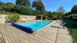 Une piscine rectangulaire avec une terrasse en bois, entourée de chaises longues et d’un mur en pierre.