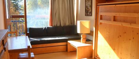 A cozy room with a wooden desk, a cushioned bench, a lamp, and a large window with curtains.