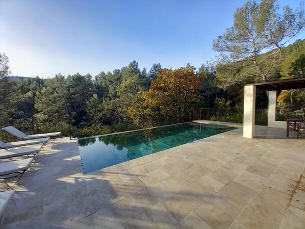 A swimming pool with lounge chairs and a pergola, surrounded by trees and hills.