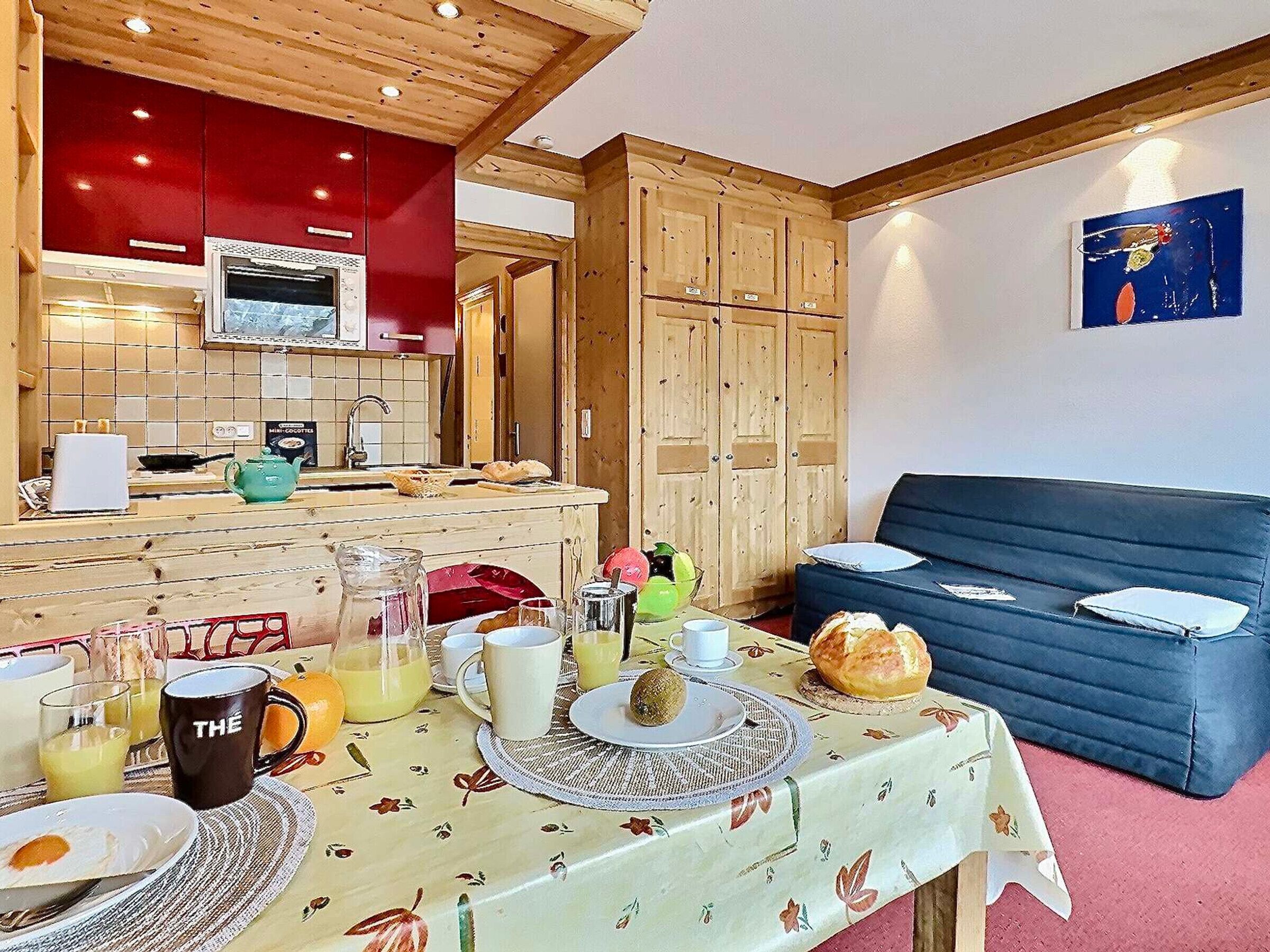 A cozy room with a dining table set for breakfast, a sofa, and a kitchen area with wooden cabinets and a red wall.
