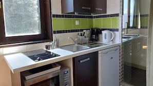 Ett kompakt kök med diskho, spis och diskmaskin. Det har ett fönster med utsikt över träd och en kakelbeklädd vägg bakom diskbänken.