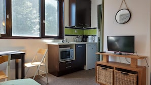 Ett kompakt kök med mikrovågsugn, ugn och kaffebryggare, en liten matplats med ett bord och en stol samt en TV placerad på ett träställ.