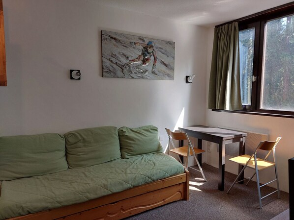 A room with a green sofa, a small table with two chairs, a window with curtains, and a painting on the wall.