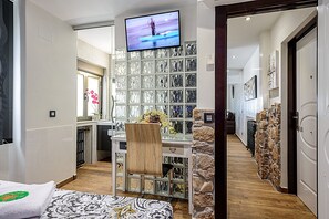 Une chambre d’hôtel avec une grande télévision, un lit recouvert d’une courtepointe à motifs, une chaise, un porte-chaussures et un mur en pierre.