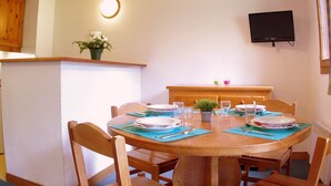 A dining area with a round wooden table set for four, surrounded by wooden chairs. There is a small potted plant on a shelf above the table, a flat-screen TV mounted on the wall, and a ceiling light fixture. - Spacious apartment with terrace, 2 bedrooms, pets allowed in Valmorel (Valmorel)