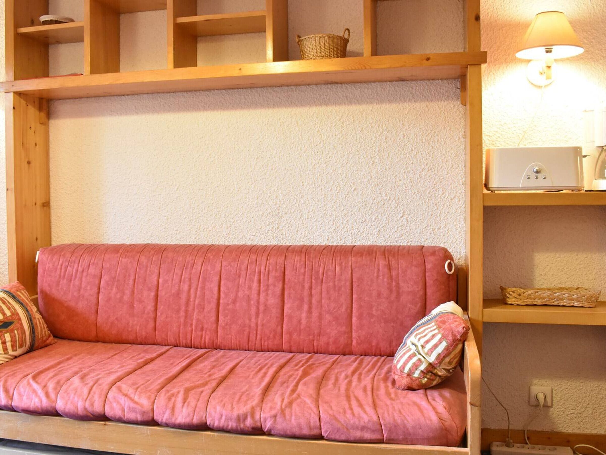 A red sofa with a cushion, wooden shelves above, and a lamp on the right.