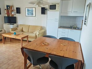 A compact living space with a sofa, a wooden dining table with two chairs, a television, a microwave, and a kitchenette. - Cozy T2 Apartment with Cabine, Pool & Fitness Access, Sleeps 6 - Le Barcarès (Port Barcarès)