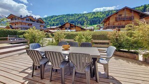 An outdoor wooden deck with a table and chairs, surrounded by lush greenery and mountain views.