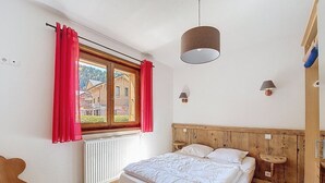 A bedroom with a wooden headboard, a bed with white bedding, a window with red curtains, and a view of houses outside.