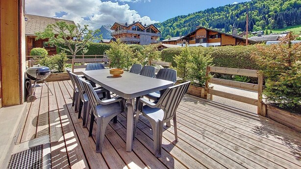 A wooden deck with a table and chairs, surrounded by greenery and overlooking a mountainous landscape.