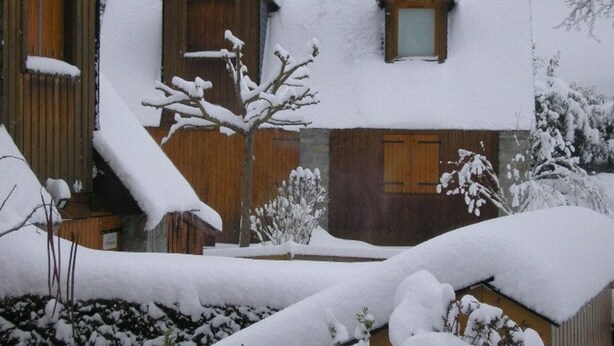A wooden cabin with a steeply pitched roof and dormer windows, surrounded by snow-covered trees and bushes.