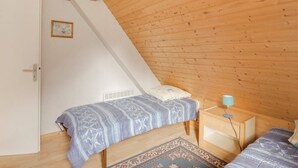 A small, cozy room with wooden walls and a sloped ceiling. There are two beds with patterned bedding, a small bedside table with a lamp, and a door with a framed picture.