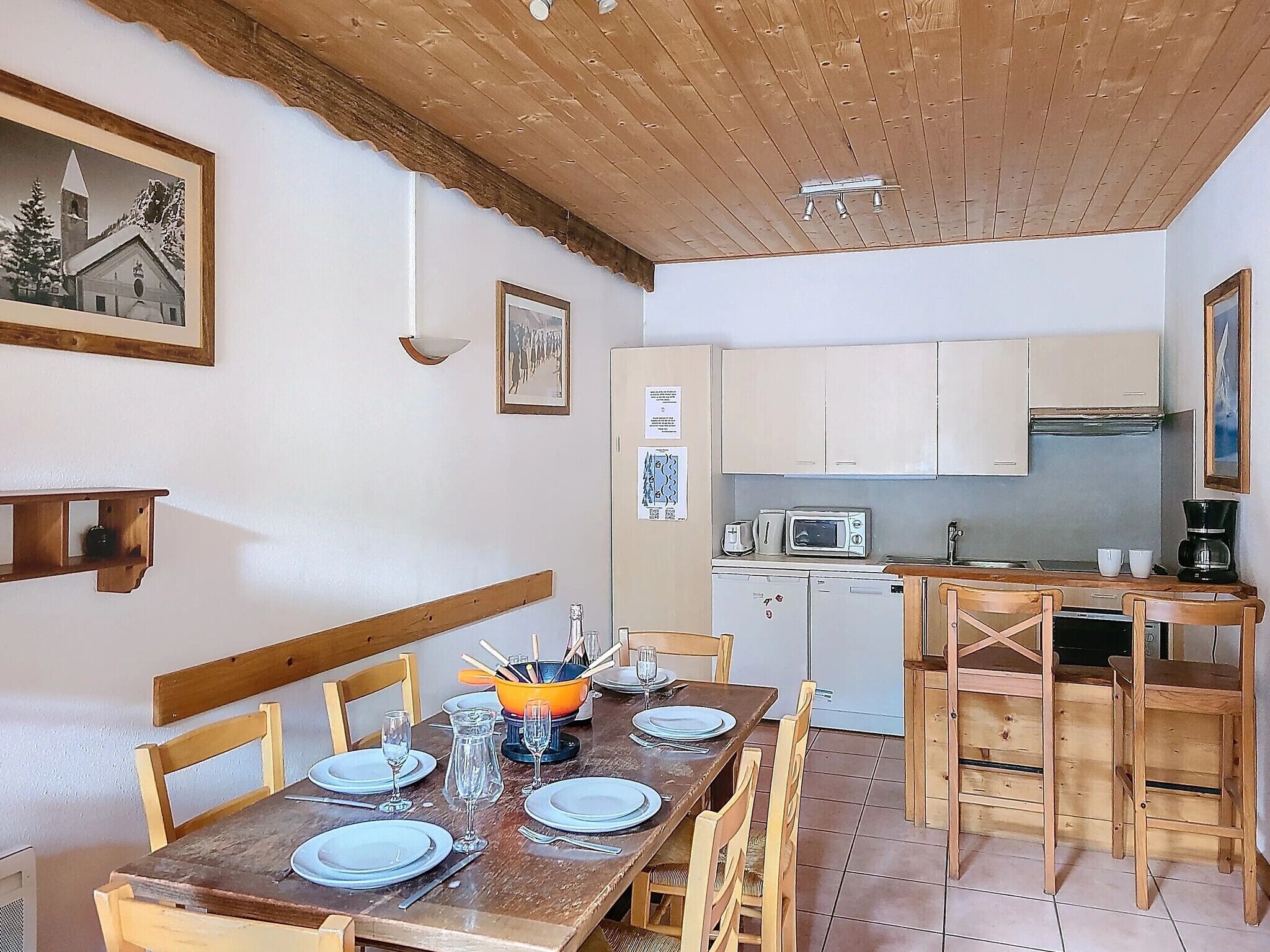 Un coin repas avec une table en bois dressée pour quatre personnes, une cuisine équipée d’appareils électroménagers de base et un plafond en bois.