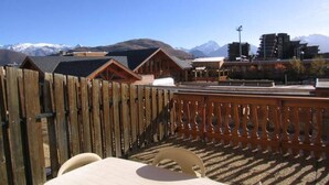 A wooden deck with a table and chairs, overlooking a mountainous landscape with buildings in the background.
