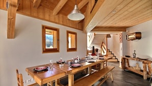 Un espace de salle à manger en bois, avec une longue table dressée pour un repas, des bancs en bois et un escalier menant à un niveau supérieur.