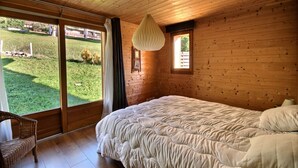 Une chambre avec un grand lit, un luminaire suspendu, une fenêtre donnant sur une cour verdoyante et un fauteuil en osier.