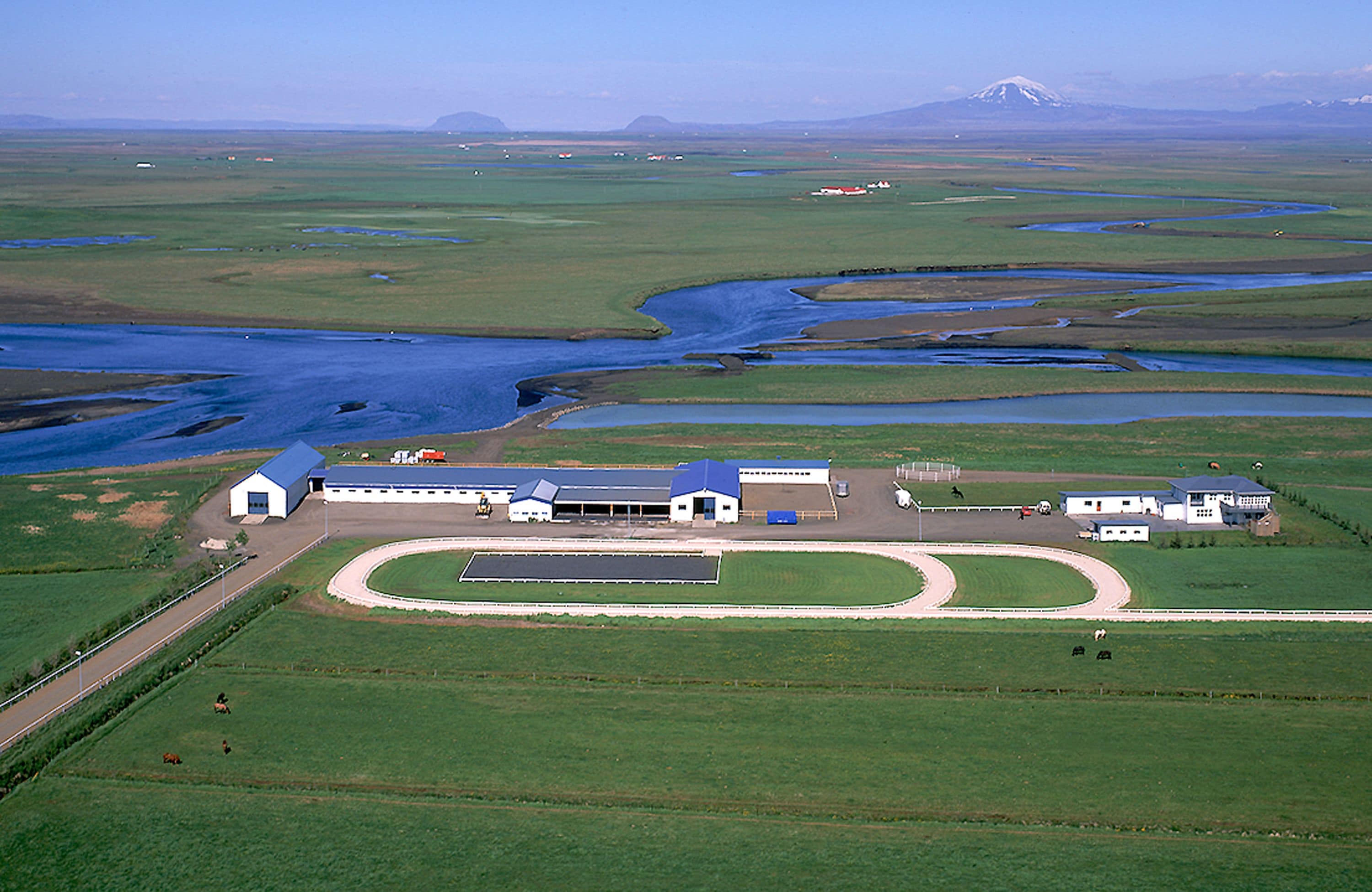 Un grand bâtiment d’un seul étage, doté d’un toit bleu, entouré de champs verdoyants et bordé d’un cours d’eau sinueux.
