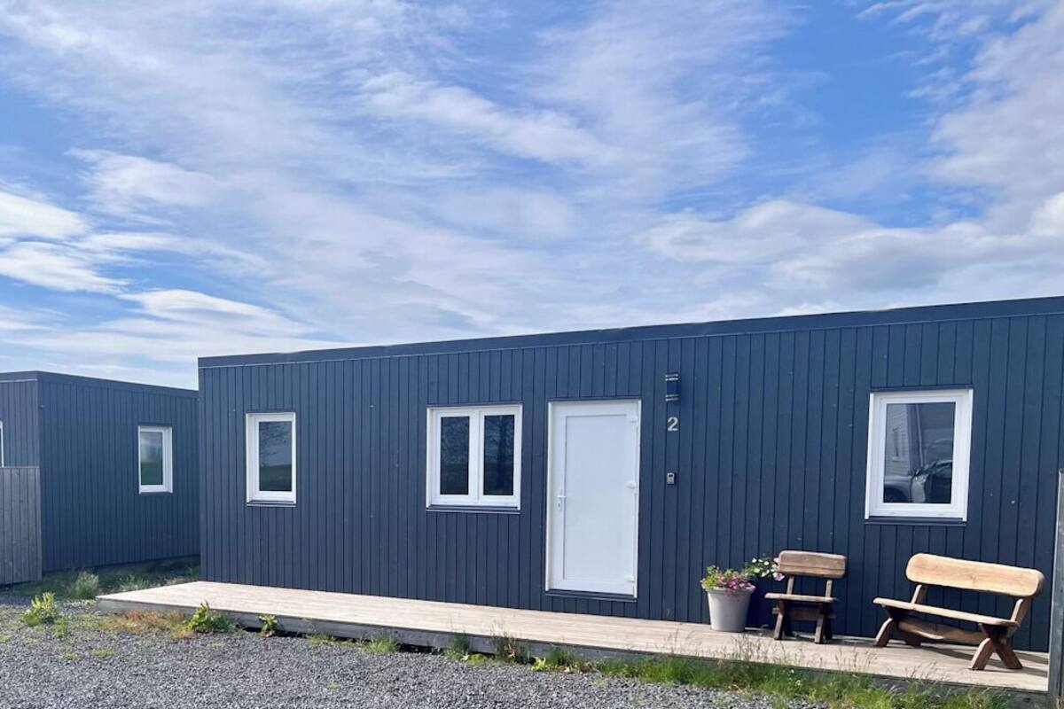 Une rangée de cabanes modernes aux couleurs sombres, dotées de fenêtres et de portes à cadre blanc, un sol recouvert de gravier et un ciel dégagé.