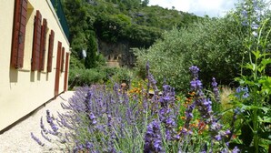 Un bâtiment à la façade jaune et aux volets rouges, entouré d’un jardin où poussent de la lavande et d’autres fleurs, avec une colline rocheuse en arrière-plan.