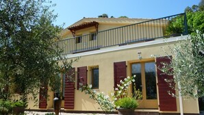 Une maison à deux étages dotée d’un balcon, de volets rouges et d’un jardin où poussent différentes espèces de plantes.