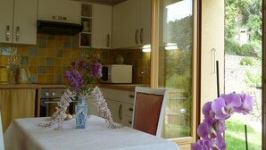 Une cuisine avec une table à manger dressée pour deux personnes, un vase rempli de fleurs et une vue sur un jardin, visible à travers une porte vitrée.