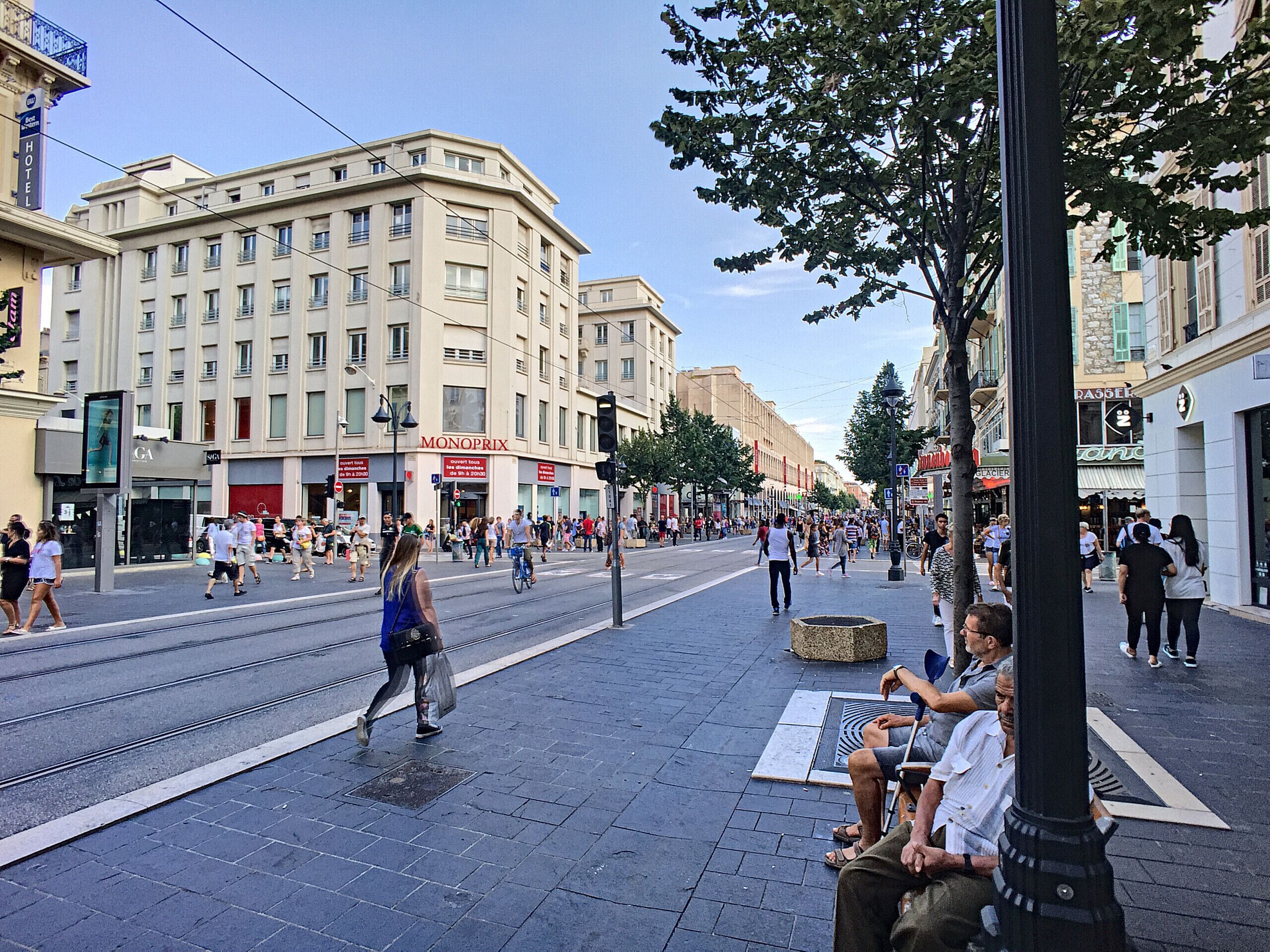 A busy urban street with tram tracks, pedestrians, and various commercial buildings including a hotel.