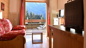 Una stanza accogliente con soffitto in legno, un divano con cuscini rossi, un piccolo tavolino, una televisione e vista sulle montagne da un balcone.