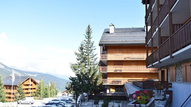 Un edificio in legno a più piani con balconi, un'area di parcheggio con auto e montagne sullo sfondo.