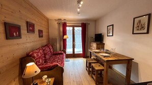 Una stanza accogliente con pavimento in legno, un divano rosso con motivo decorativo, un piccolo tavolo in legno con una lampada, un televisore su un mobiletto in legno e un tavolo da pranzo per due persone.