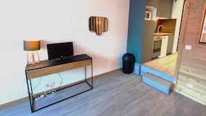 Ein modernes, kompaktes Zimmer mit Schreibtisch aus Holz, Fernseher und einer Wandleuchte. Man sieht einen Teil einer Küche.